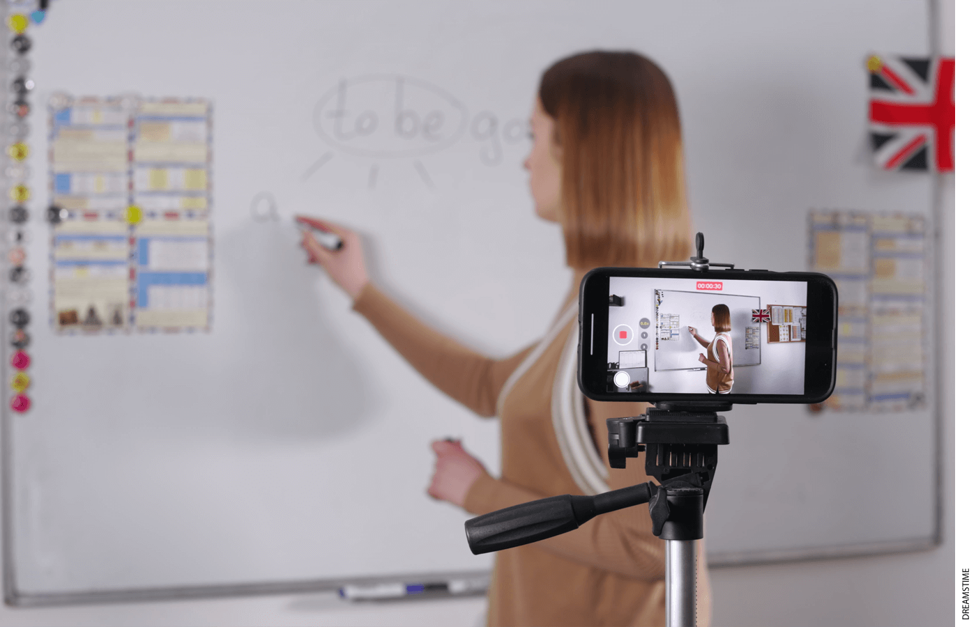 Photo of a teacher writing on a white board while being filmed on a phone